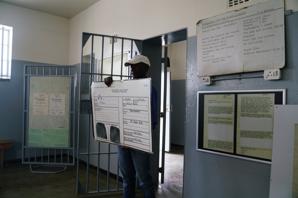 Vusi Kube mit einem Robben-Island-Gefangenenausweis (nicht seinem eigenen), den die Gefangenen eigentlich immer dabei haben mussten. An der Wand die Tagesrationen und ein zensierter Brief.