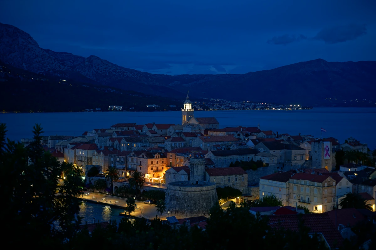 Korčula-Stadt (Stari Grad – „Altstadt“) in der Blauen Stunde, gesehen von der Stichstraße links neben dem Aussichtspunkt mit dem kleinen weißen Korčula-Schriftzug; im Hintergrund Orebić auf dem „Festland“
