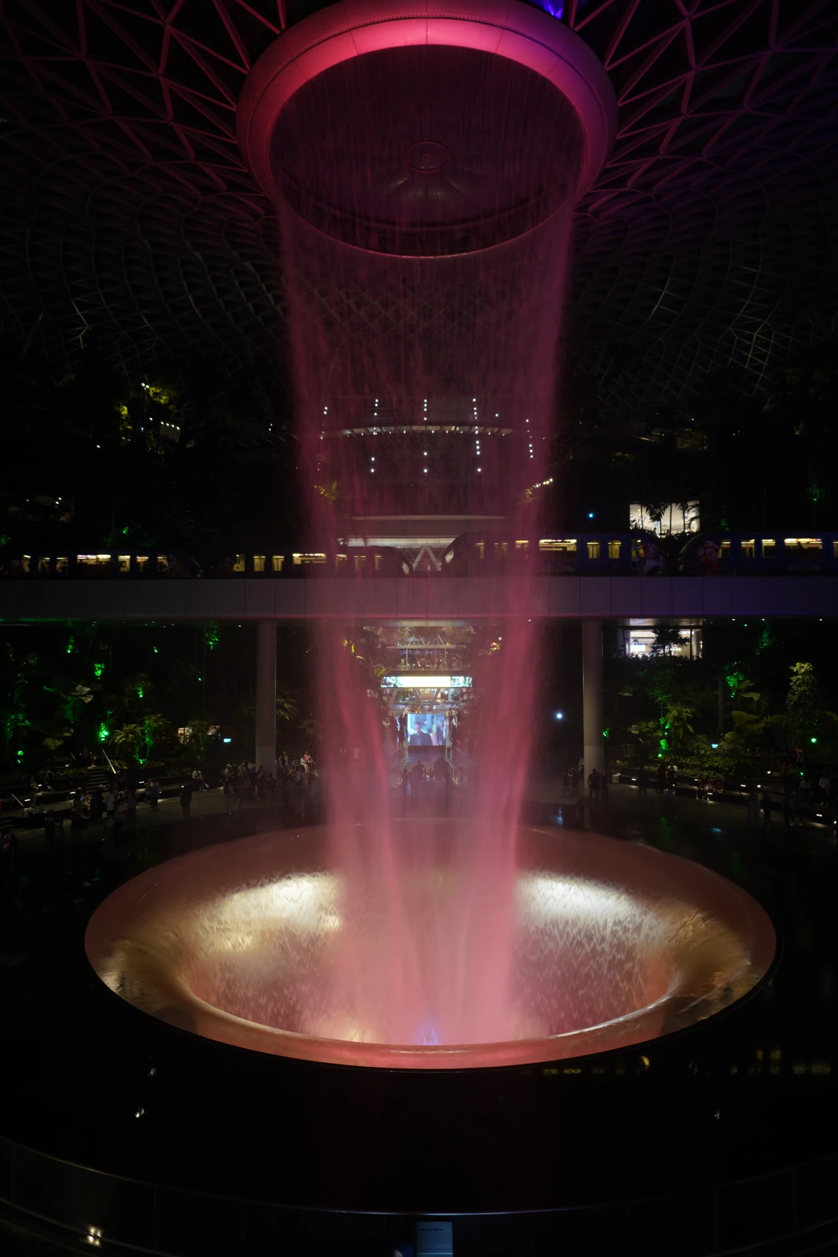 Rain Vortex im Jewel Changi Airport in Singapur bei Nacht, im Hintergrund zwei Züge